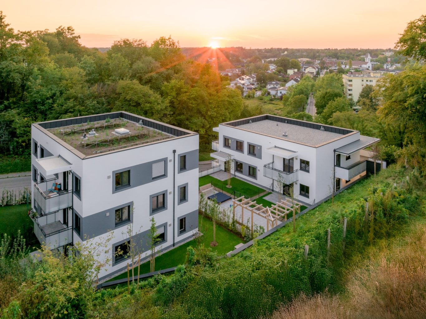 Gauting Zwei moderne Wohngebäude mit Terrasse, umgeben von Grün und einem Sonnenuntergang im Hintergrund.